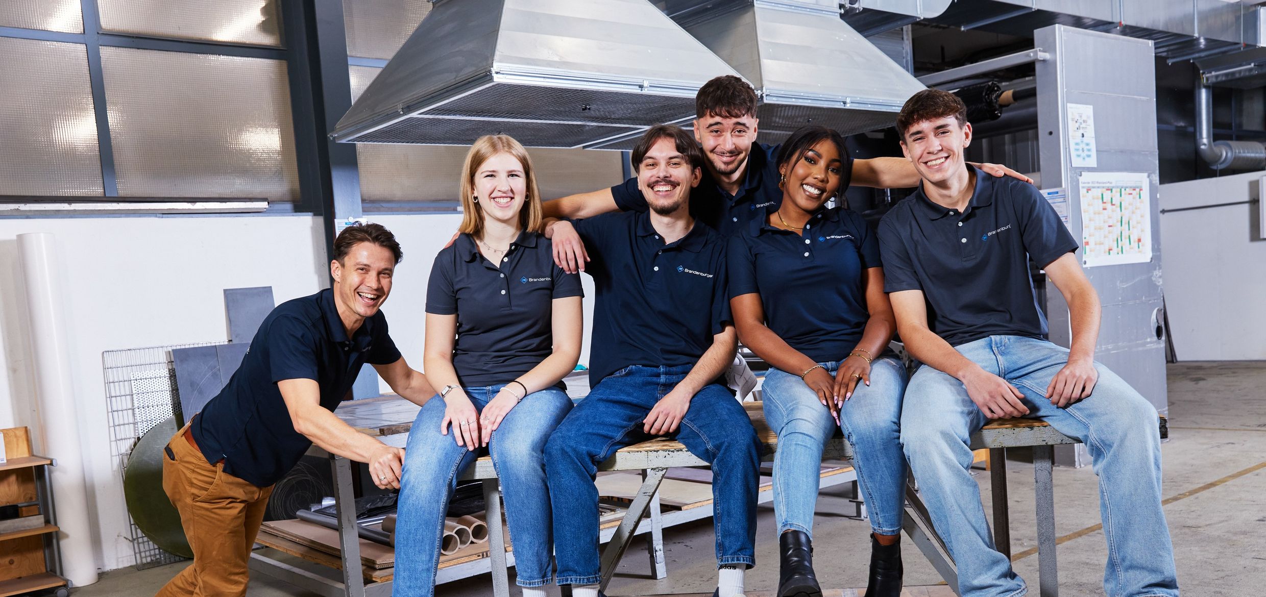 A group of six smiling people in casual attire, sitting and standing in an industrial setting with large vents and equipment in the background. A group of six smiling people in casual attire, sitting and standing in an industrial setting with large vents and equipment in the background.