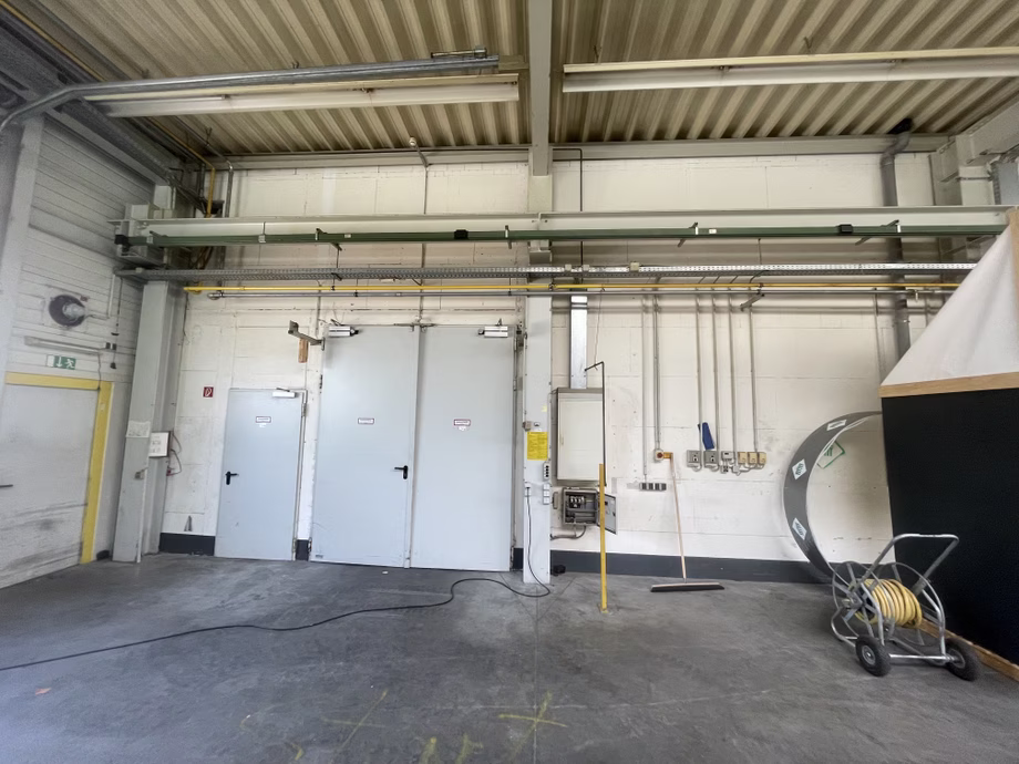Industrial interior with two gray doors, overhead pipes, a hose reel, and a concrete floor. Walls have electrical panels and wiring.
