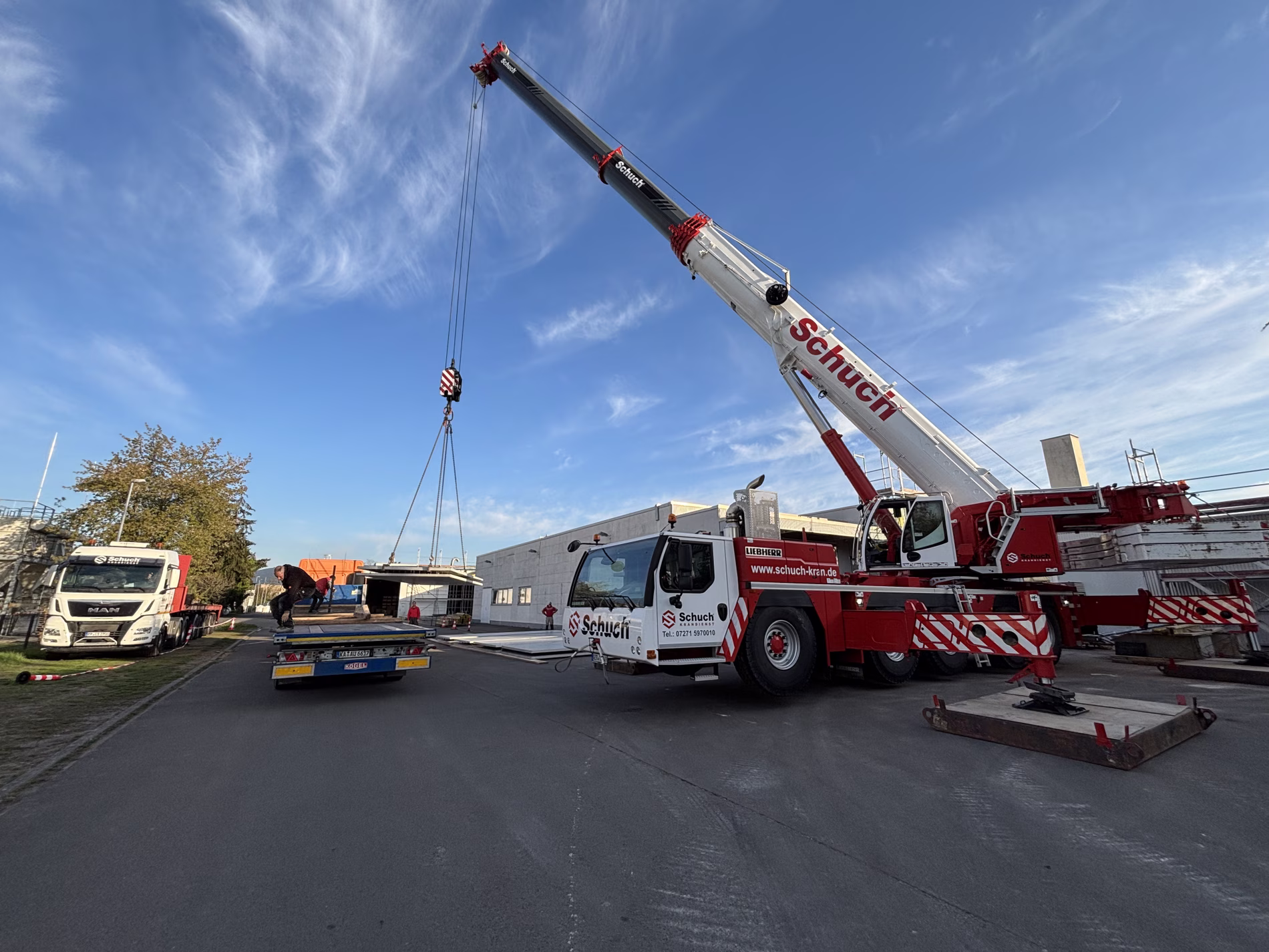 A large crane with "Schuch" branding lifts materials at a construction site. Trucks are parked nearby under a clear blue sky.