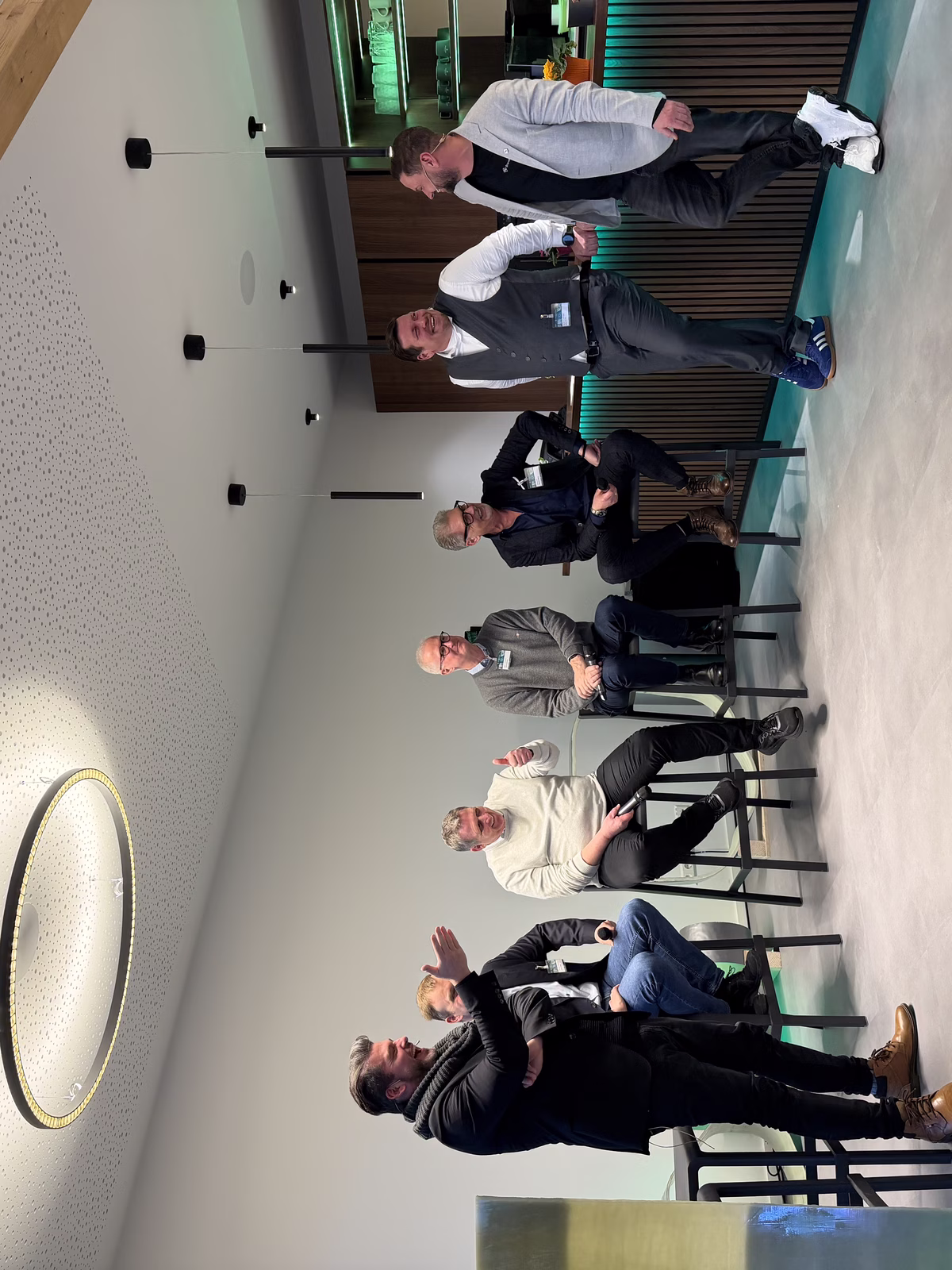 A group of men are casually talking and sitting on stools in a modern room with a bar, under a circular ceiling light.