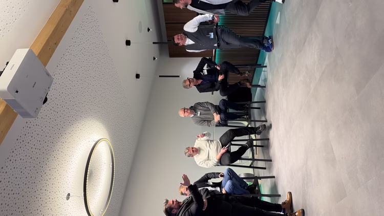 A group of men are casually talking and sitting on stools in a modern room with a bar, under a circular ceiling light.