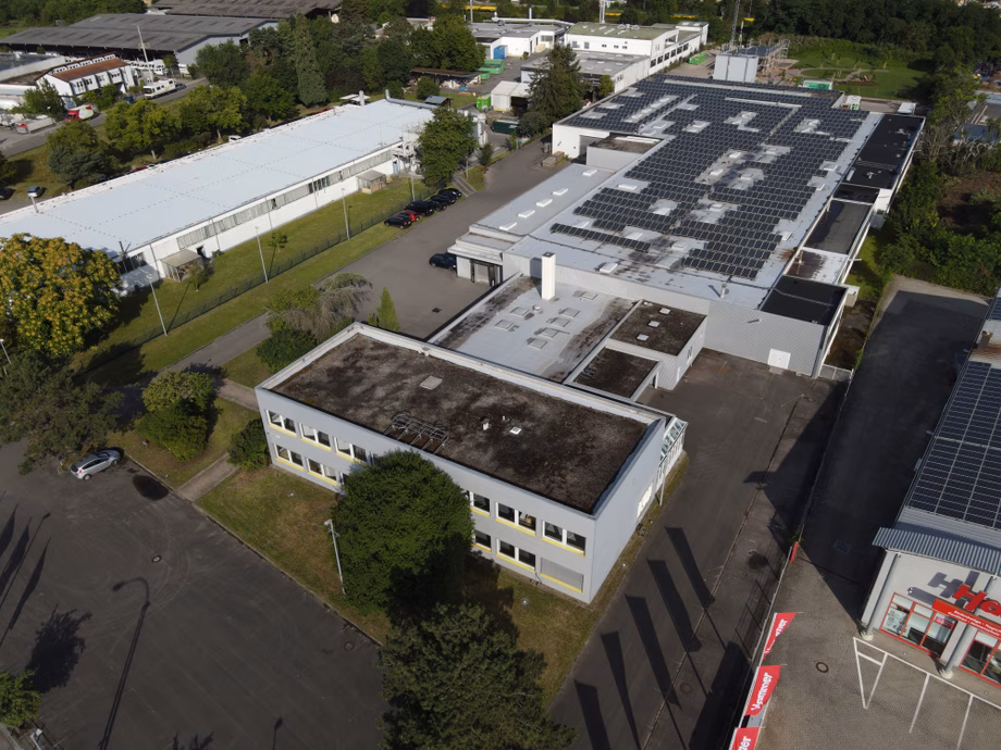 Aerial view of an industrial complex with buildings featuring solar panels on the roofs, surrounded by trees and roads.