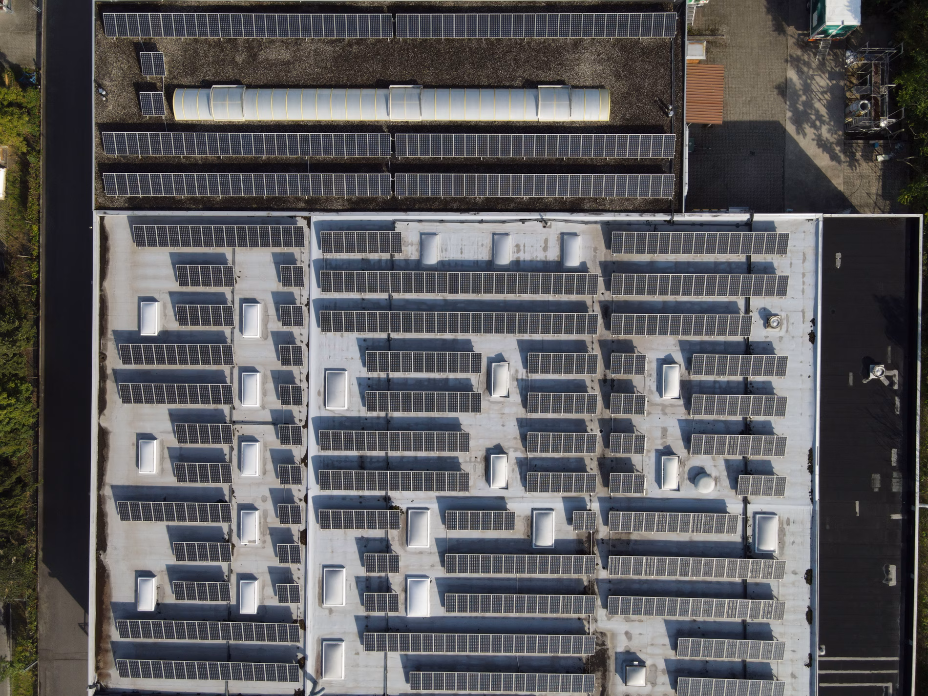 Aerial view of a large rooftop covered with numerous solar panels, arranged in rows, surrounded by greenery and industrial structures.