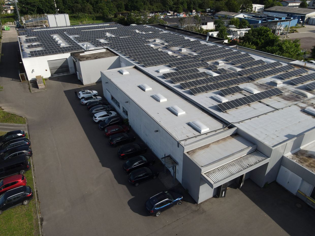 Aerial view of a large industrial building with solar panels on the roof and parked cars along the side in a suburban area.