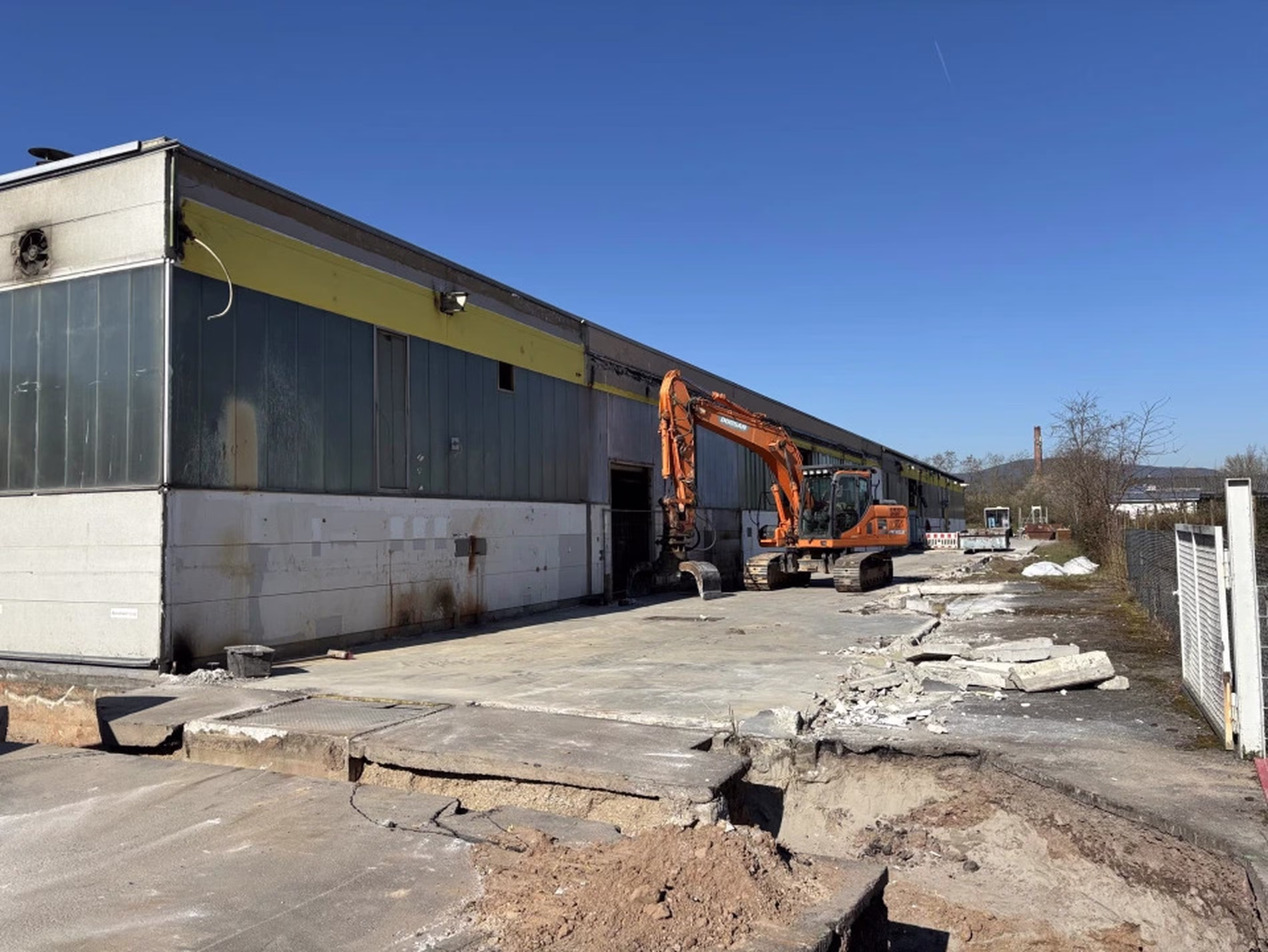 An orange excavator demolishes a section of a large industrial building under a clear blue sky, with debris scattered on the ground.