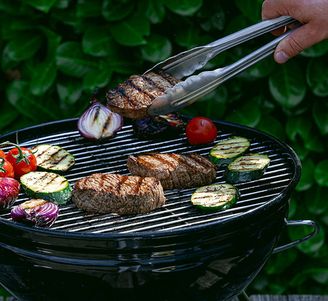Hand holding tongs flips grilled steak on a barbecue with zucchini, tomatoes, and onions against green foliage.