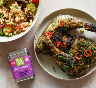Plate of herb‑rubbed grilled chicken legs with sliced chilies, beside a bowl of couscous salad and a tin of Ras el Hanout spice.