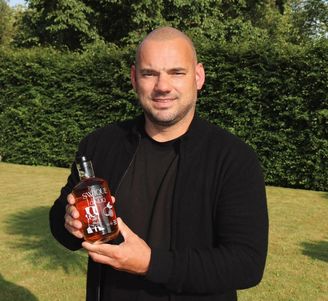 Bald man in black cardigan smiling outdoors, holding a bottle of amber liquor with trees and grass in the background.