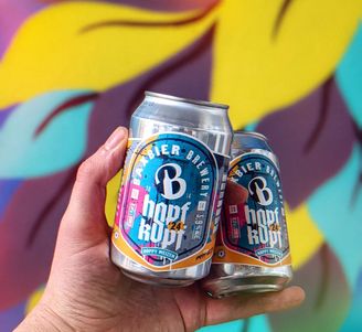 Hand holding two colorful craft beer cans against a bright, abstract yellow and purple mural background.
