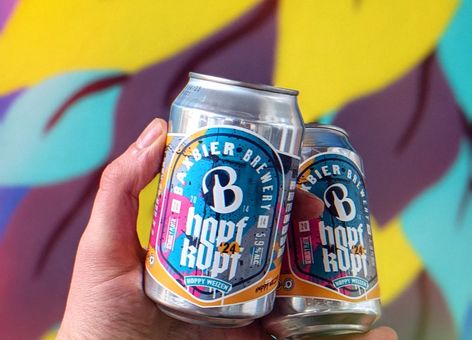 Hand holding two colorful craft beer cans against a bright, abstract yellow and purple mural background.