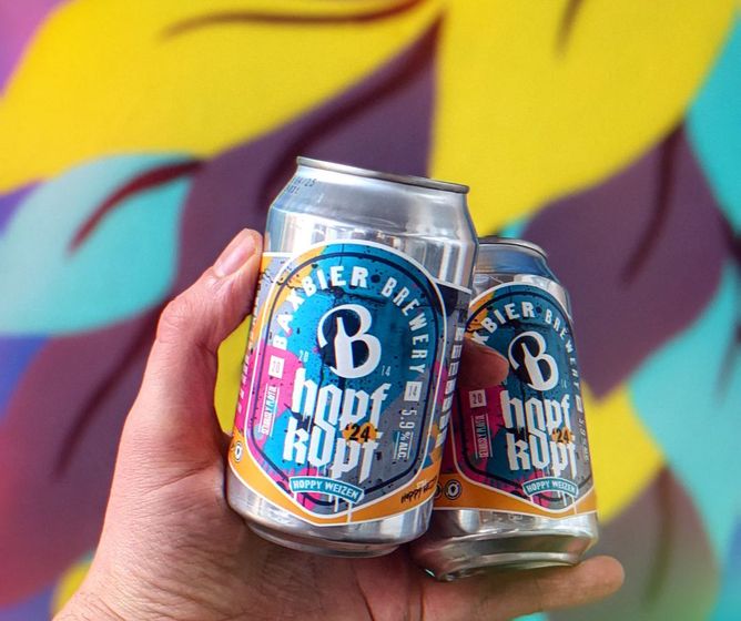 Hand holding two colorful craft beer cans against a bright, abstract yellow and purple mural background.