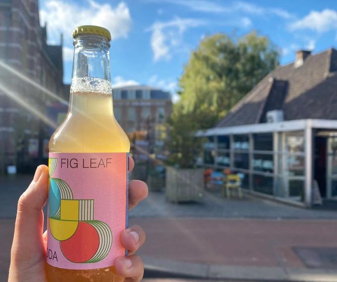 Hand holding a colorful fig leaf soda bottle on a sunny street with buildings and outdoor seating in the background.
