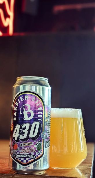 Tall Baxbier Brewery 430 IPA can beside a hazy golden beer in a stemless glass on a bar table, neon lights blurred in background.