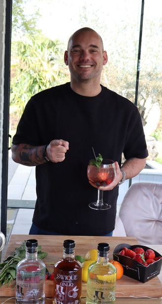 Smiling man indoors holds a red cocktail behind a counter with gin bottles, herbs, citrus fruits, and fresh strawberries.