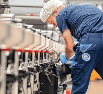 Een werknemer in een blauw uniform en een haarnetje stelt machines af in een fabriek, waarbij hij zijn aandacht richt op een zwart onderdeel langs de productielijn.