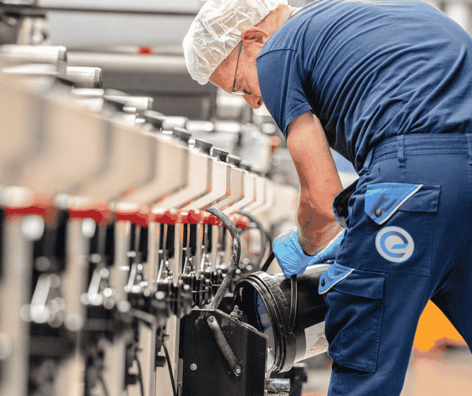 Een werknemer in een blauw uniform en een haarnetje stelt machines af in een fabriek, waarbij hij zijn aandacht richt op een zwart onderdeel langs de productielijn.