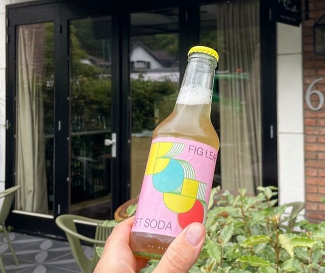 Hand holding a colorful fig leaf soft soda bottle in front of a modern café with glass doors and greenery.BOJOCraftSoda_sfeer