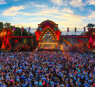 Huge outdoor music festival crowd facing a colorful, illuminated main stage at sunset under a dramatic sky.