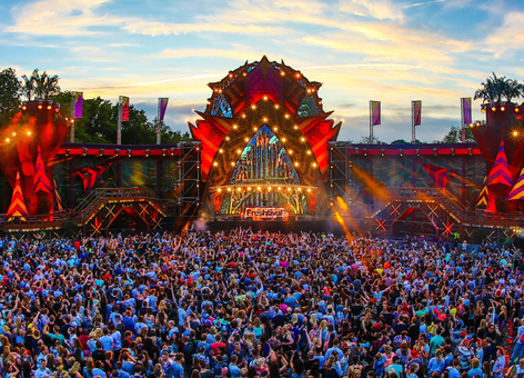 Huge outdoor music festival crowd facing a colorful, illuminated main stage at sunset under a dramatic sky.