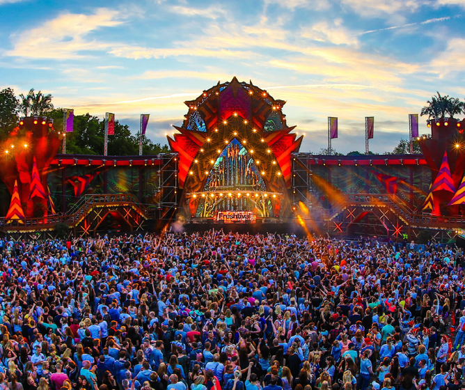 Huge outdoor music festival crowd facing a colorful, illuminated main stage at sunset under a dramatic sky.