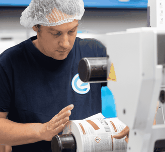 Een medewerker met een haarnetje bedient een etiketteermachine en brengt waarschuwingsetiketten voor chemische stoffen aan op een rol in een productiefaciliteit.