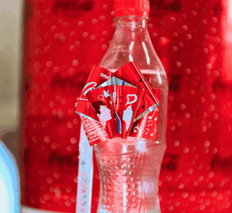 Een doorzichtige Coca-Cola-fles met rode etiketten met hartjesmotief tegen een wazige rode Coca-Cola-achtergrond.