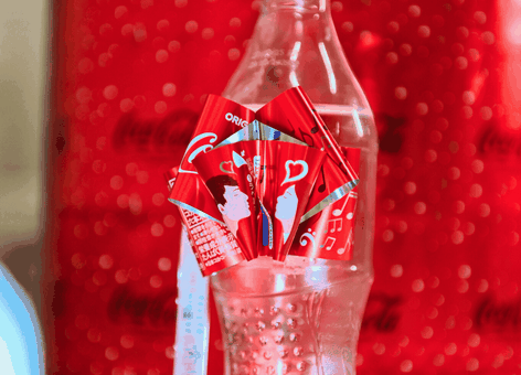 Een doorzichtige Coca-Cola-fles met rode etiketten met hartjesmotief tegen een wazige rode Coca-Cola-achtergrond.