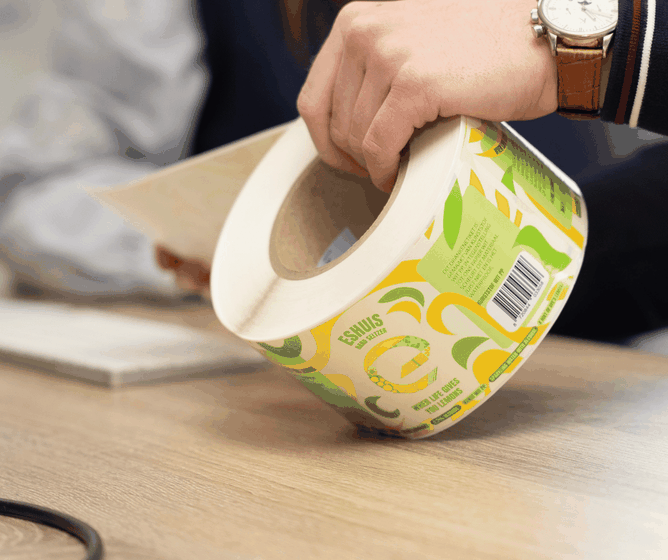 Person peeling a bright green and yellow product label from a large roll on a wooden desk.