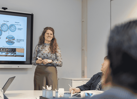 Woman giving a presentation beside a screen with technical diagrams, speaking to seated colleagues in a meeting room.