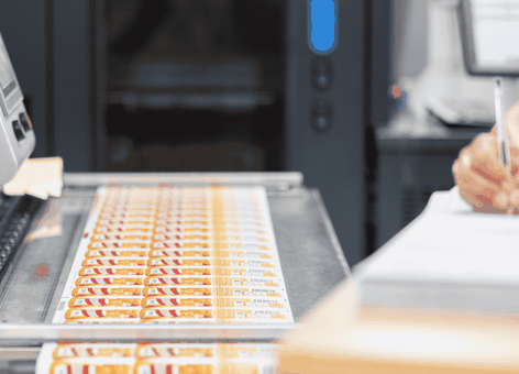 Color labels emerging from an industrial printer while a person beside it writes notes on a clipboard.
