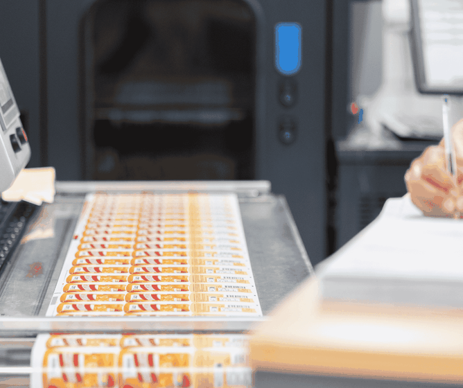 Color labels emerging from an industrial printer while a person beside it writes notes on a clipboard.