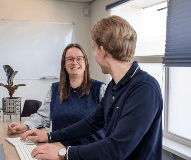 Two colleagues talking at a desk by a window, one typing on a keyboard while the other smiles and listens.
