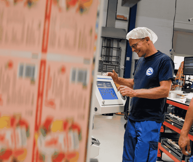 Een medewerker in een blauw uniform en een haarnetje die glimlacht terwijl hij een touchscreenmachine bedient in een drukkerij..