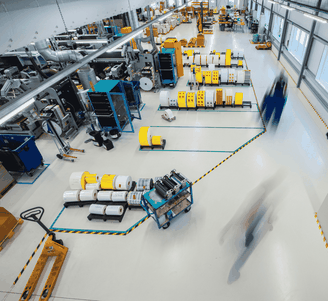 Een breed beeld van een lichte fabrieksvloer met machines, grote spoelen en vage silhouetten van werknemers die zich tussen de overzichtelijke werkplekken bewegen.
