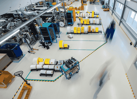 Een breed beeld van een lichte fabrieksvloer met machines, grote spoelen en vage silhouetten van werknemers die zich tussen de overzichtelijke werkplekken bewegen.
