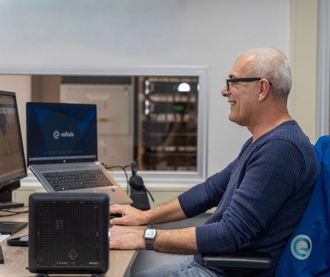 Smiling man with glasses works at a desk with two computer monitors in an office, wearing a smartwatch and blue long-sleeve shirt.