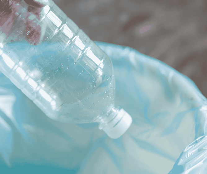 Hand dropping an empty plastic bottle into a blue plastic-lined trash or recycling bin.