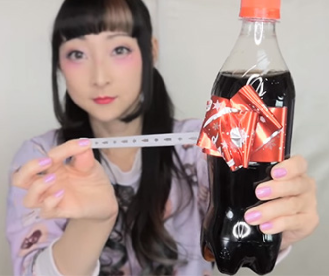 Japanse vrouw kijkt naar de camera, terwijl ze in de ene hand een versierde frisdrankfles en in de andere een strookje papier vasthoudt.