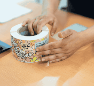Handen rollen een kleurrijke rol met bloemenetiketten van „Yaya Kombucha“ uit op een houten bureau.