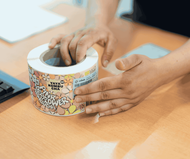 Handen rollen een kleurrijke rol met bloemenetiketten van „Yaya Kombucha“ uit op een houten bureau.