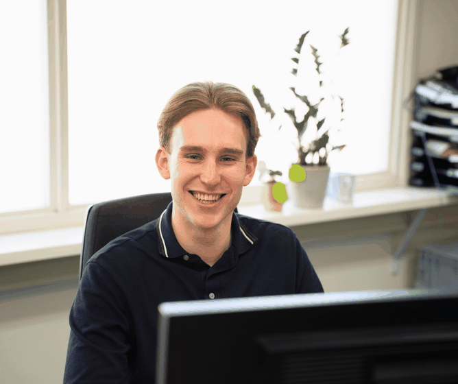 Smiling young man in a navy polo shirt sitting at a desk, working at a computer in a bright office with a potted plant in the background.