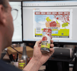 Person comparing a printed apple juice can with its digital label design on a computer screen in an office setting.