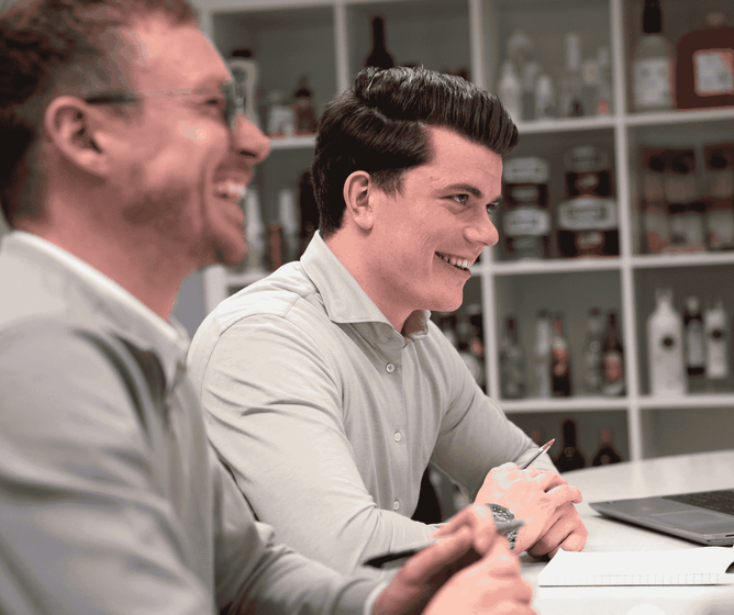 Two smiling men in a meeting sit at a table with notebooks and a laptop, shelves of bottles blurred in the background.