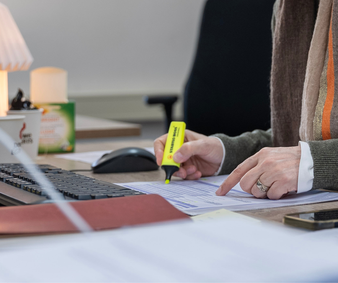 Person at a desk highlighting text on a printed document beside a keyboard, mouse, coffee cup, and smartphone.