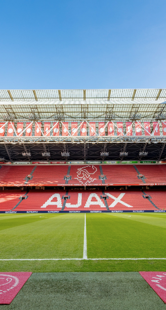 Empty Johan Cruyff Arena football stadium with green pitch, red Ajax seating, and retractable roof under a clear blue sky.