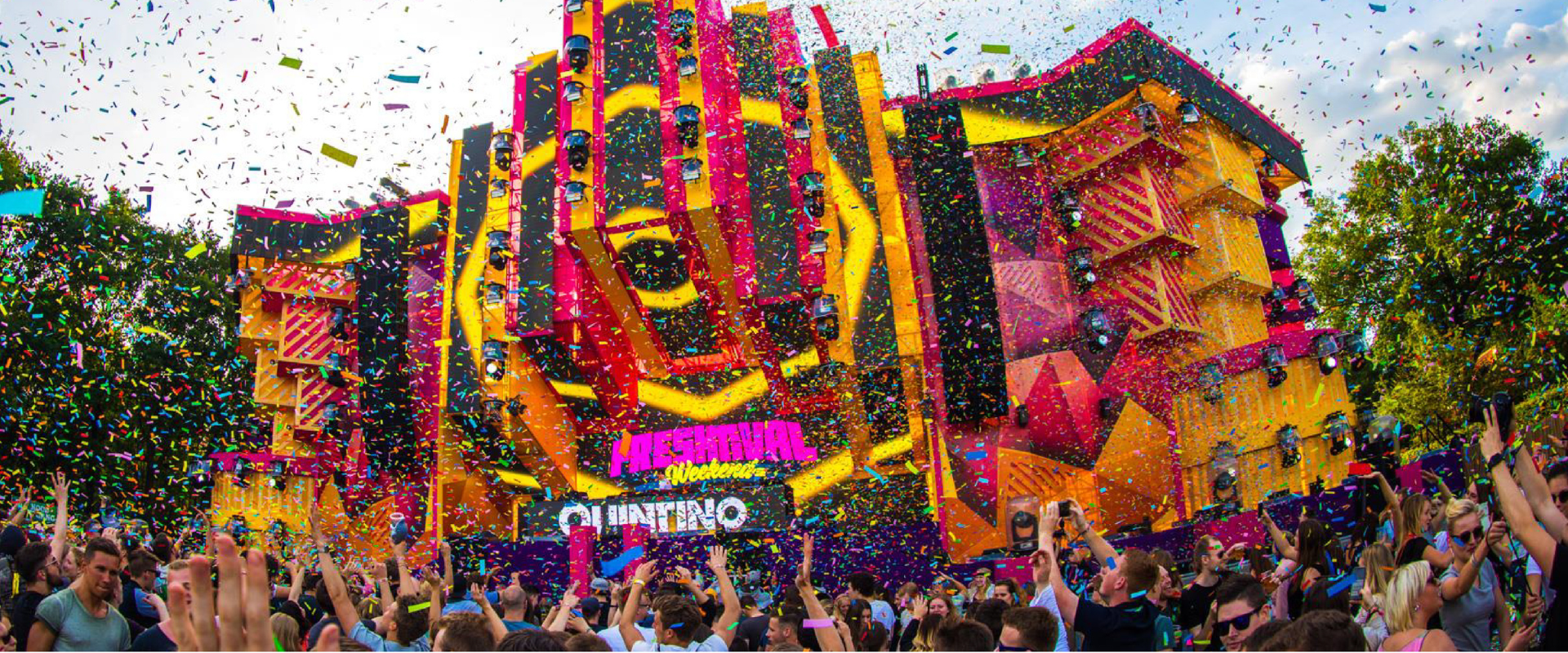 Large crowd partying amidst colorful confetti in front of a bright, geometric outdoor festival stage.