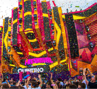 Large crowd partying amidst colorful confetti in front of a bright, geometric outdoor festival stage.