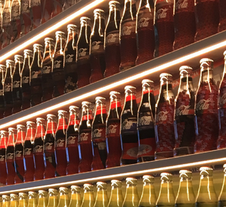 Rows of illuminated glass soda bottles lined up on angled shelves, creating a colorful, repeating pattern.