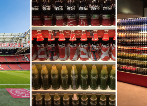 Empty Johan Cruyff Arena football stadium with green pitch, red Ajax seating, and retractable roof under a clear blue sky.