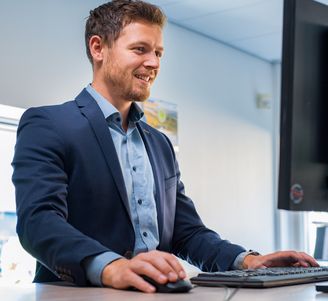 Man in a blue suit using a computer at a standing desk, smiling, in a bright office environment.